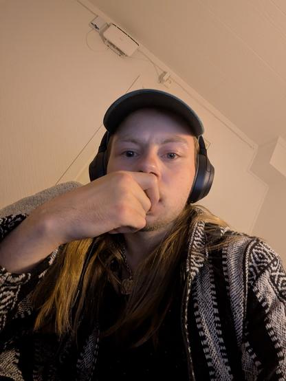 Young man wearing a green cap, headphones, and a patterned jacket, looking directly at the camera, hand near mouth.
