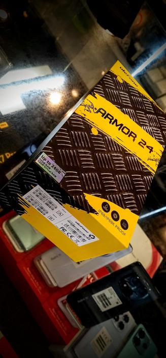 The photograph shows a product box for a smartphone, prominently featuring the text "ARMOR 24" in bold, black letters on a yellow background. The box has a textured brown surface with a diamond plate pattern. A sticker on the box indicates "Up to 24GB RAM" and "12GB Virtual RAM." Below this, there is a label with a barcode and additional text, including "Model: Armor 24" and "IP68/IP69K PROOF," suggesting the phone's water and dust resistance. The box is placed on a reflective black surface, with other phone boxes visible in the background, including a red one and a black one with a camera module visible. The lighting in the image is dim, with reflections visible on the surface.