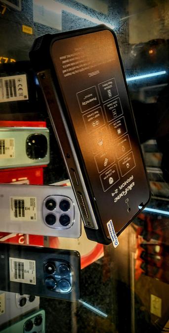 The photograph features a massive black Android phone  with the brand name "Ulefone" and the model "ARMOR 24" prominently displayed on its back. The Android is designed with a textured surface and includes a set of icons and text, likely indicating various functions or features. The phone is positioned at an angle, showcasing its side profile, which includes a charging port and a headphone jack.

In the background, several smartphones are visible, each with a different color and camera setup. The phones are placed on a reflective surface, creating a mirrored effect. The colors of the phones include light blue, red, and dark blue, with each phone featuring a camera module with multiple lenses. The setting appears to be a retail environment, with a glass display case and a dimly lit background, enhancing the focus on the products.
