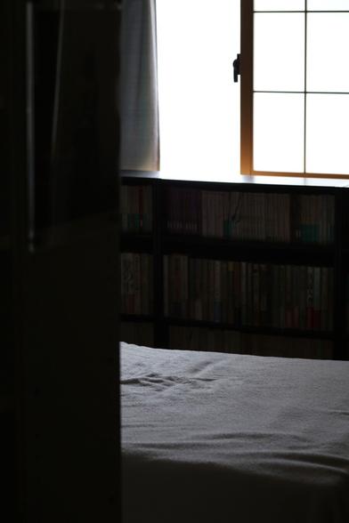 The bed room seen from the hall next to it. It's bright outside the window but dim inside. The white blanket  covering the bed reflects the light filtering through from outside.