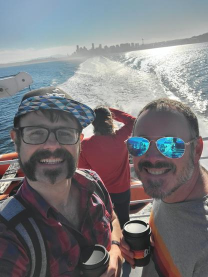 A selfie with my husband. We're on a ferry and Seattle is behind us. You can see a large wake from the ferry. My husband has shiny refective glasses and you can see my arm holding up the phone in the reflection. We're both holding coffees.
