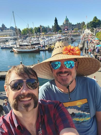 A selfie of my husband and me on the Victoria harbor. There are large crowds behind us gathered for Canada day. In the distance is a big temporary stage erected in front of a civic building. A large Canadian flag is hanging from the building.