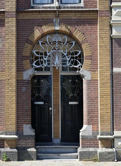 Double porte insérée dans un renfoncement en brique. Une joyeuse alternance de briques rouges et jaunes en habille le tour. Le haut forme un peu plus qu'un demi-cercle un peu moins qu'un cercle complet, très élégant, dont le centre est occupé par des motifs de ferronnerie abstraits Art nouveau très sophistiqués. Il y a aussi des volutes décoratives sur chacune des portes.