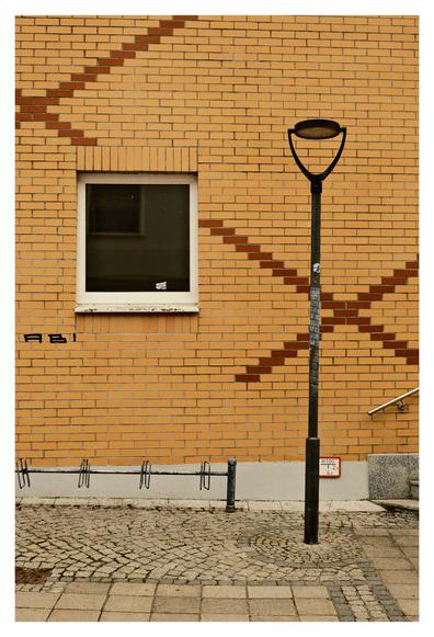 Eine gelbe Backsteinwand mit diagonalen Mustern aus dunkleren Ziegeln. Vor der Wand steht eine schwarze Straßenlaterne. Links ist ein kleines Fenster mit einem Aufkleber und Abplatzungen am Rahmen zu sehen. Unten verläuft ein Fahrradständer auf einem gepflasterten Gehweg, rechts führen Stufen mit einem Edelstahlgeländer hinauf. An der Laterne ist ein kleines Schild mit Hydrantenangabe befestigt.