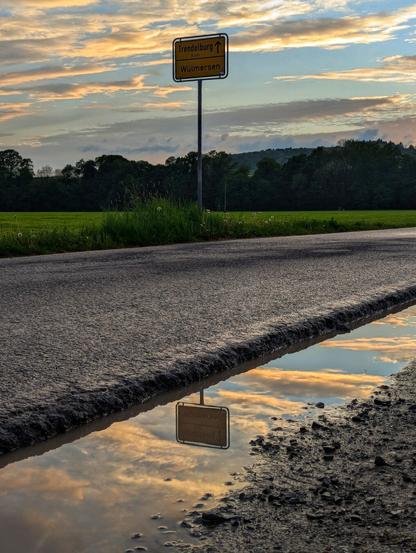 A low-angle shot captures a road stretching into the distance with a puddle reflecting the sky in the foreground. The sky is filled with dramatic, illuminated clouds at what appears to be either sunrise or sunset. A yellow signpost with black text indicates directions to "Trendelburg" and "Wülmersen". Green fields line the left side of the road, and a dark, dense forest stands on the horizon under the vibrant sky. The reflection of the sky and the signpost is visible in the muddy puddle next to the road.