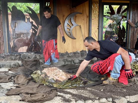 A photo of two men removing a whole pig from its underground oven.