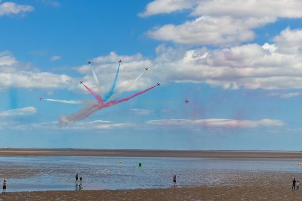 This photograph captures an airshow performance by the Red Arrows, the Royal Air Force’s aerobatic team. Against a backdrop of a soft blue sky with scattered clouds, nine aircraft soar upward in a perfect fan-shaped formation, trailing vivid streams of red, white, and blue smoke. The smoke trails radiate symmetrically like the petals of a flower opening in midair. Below, on a reflective wet beach, small figures of people stand still, gazing upward.