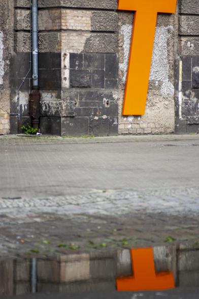 Eine alte Hausfassade, an dem ein grosses orangenes T hängt, das sich in einer Pfütze davor spiegelt.