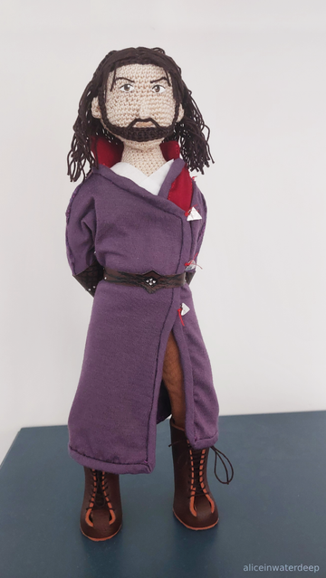 Front view of a handmade Gale of Waterdeep doll with his hands behind his back, standing on a blue surface. The doll features a crocheted beige body with embroidered facial features, shoulder-length brown yarn hair, and wears a purple wizard robe with red collar, brown leather belt and bracers with silver studs, and laced brown boots.