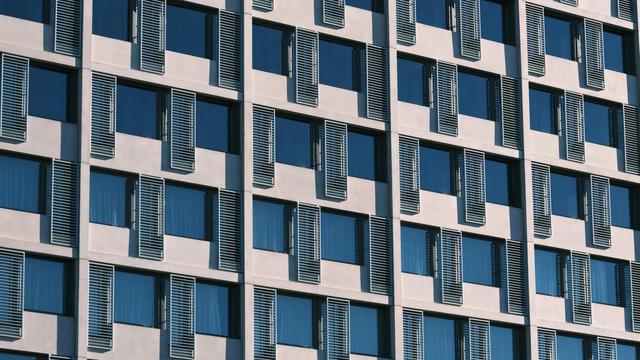A photo of a windowed wall with an array of three different elements. Smaller square blue windows, reflecting the sky. Longer and narrow metal meshes inbetween, framed by white concrete for the structure