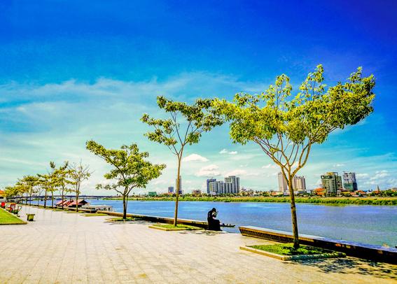 Photo shows a brick laid square and sidewalk to a wide blue river. Low rails of brickwork made for sitting border the walking area from the stairs down to the river. Along the sidewalk and the siderail there are low trees with large, shadowing crowns. Along the walkway to the left there are benches and bins. In the background at the left side of the river there are some house boats with red roofs visible. On the other side of the river a low area of land with mix building types and heights from two floors to high rises shows a city in development.