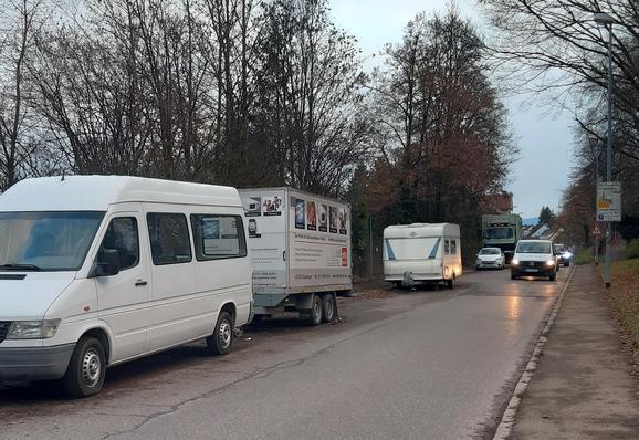 Das Bild zeigt die Sulzgrieser Straße, fotografiert aus Richtung Rüdern. Auf der linken Seite der Straße steht ein weißer Minivan mit einem Anhänger mit einer Werbeplatte. Neben dem Minivan steht ein weißer Wohnwagen mit blauen Streifen. Auf der rechten Seite fährt ein weißes Auto mit eingeschaltetem Scheinwerfer auf der Straße. Im Hintergrund sind weitere Fahrzeuge zu sehen, darunter ein grüner Lastwagen. Die Straße ist asphaltiert und von Bäumen gesäumt, die im Herbst ihre Blätter verloren haben.