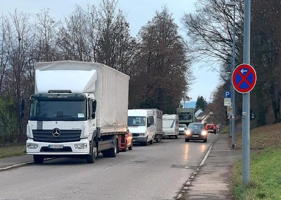 Das Bild die Sulzgrieser Straße, gesehen aus Richtung Rüdern. Auf der linken Fahrspur parken mehrere Fahrzeuge, darunter ein großes weiße Mercedes-Lkw, ein kleinerer weißer Transporter und ein Wohnwagen, in einer Reihe. Ein rotes Auto fährt in die entgegengesetzte Richtung.