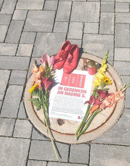 Gedenktafel Auf einem gepflasterten Boden. Flugblatt liegt dort:"Im Gedenken an Nadine S."
Rechts und links ein Blumenstrauß. Ein Grablicht sowie ein paar rote Schuhe