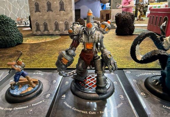 A tabletop gaming scene featuring three miniatures on a textured game board. The central figure is a large, metallic construct with glowing orange elements. To its left is a smaller figure, "KANG," depicted as a character wielding a crowbar.