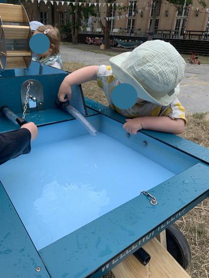 Das Bild zeigt unkenntlich Kinder, die am blauen Wellenspieler mit Wasser spielen.