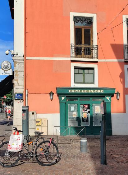 Fachada de un edificio pintada en color naranja. En ella está la puerta de un café pintada de verde. El café de llama "cafe le flore". Atada a una valla de la calle hay una bici de la que cuelga una bolsa. En medio de la calle hay una pilona.