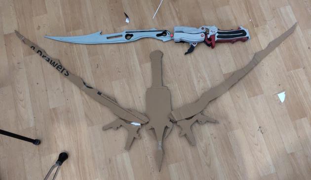 Two weapons lying on the floor. Silver fantasy sword on top, made from wood, fully painted. It's Lightning's gunblade from Final Fantasy XIII. Below is a crossbow with some odd shapes, made of cardboard. It's significantly larger than the sword. It's Serah's bow from Final Fantasy XIII-2.