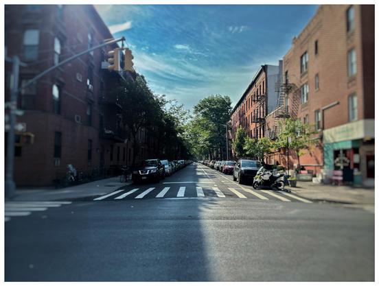 View from an intersection in a residential neighborhood. Trees and parked cars line the curbs. Apartment buildings recede to a central vanishing point. The sky is blue with high cirrus clouds. The midafternoon sun illuminates the buildings on the right side of the image while throwing the left side into shadow, with the dividing line between light and dark going right down the middle of the street.