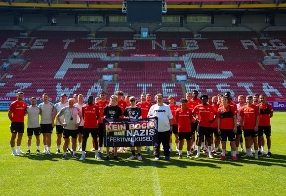 Eine Gruppe von Fußballspielern und Mitarbeitern posiert auf dem Feld eines Stadions. Sie tragen Sporttrikots, meist rot, und halten ein Banner mit der Aufschrift "KEIN BOCK auf NAZIS FESTIVAL KUSEL".