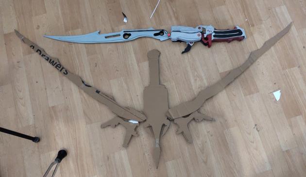 Two weapons lying on the floor. Silver fantasy sword on top, made from wood, fully painted. It's Lightning's gunblade from Final Fantasy XIII. Below is a crossbow with some odd shapes, made of cardboard. It's significantly larger than the sword. It's Serah's bow from Final Fantasy XIII-2.