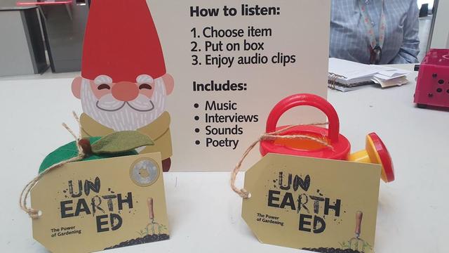 A small watering can and a green pumpkin sit on a white desk with signs saying Unearthed in front of them. Behind is a illustrated Gnome on printed card with instructions on how to enjoy MiaB.