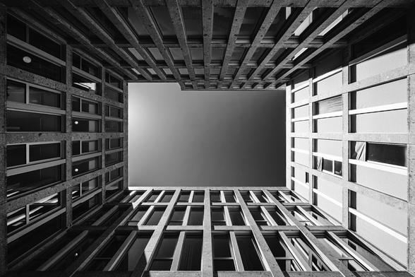 Black and white upward view of a modern square courtyard enclosed by concrete and glass buildings, with the clear sky forming a stark geometric opening at the center. The architectural grid creates strong lines and symmetry.