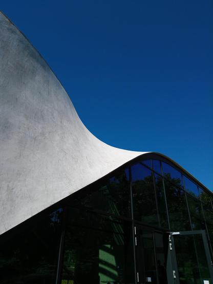 Oberhalb einer Glasfassade schwingt sich eine dünne Betonwand zum flachen Dach auf. Es ist eine dynamische Bewegung.
