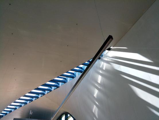 Die Decke ist lang und spitzoval durchbrochen. Die Aufnahme wurde von innen gemacht. Deckenlichter lassen das Sonnenlicht hindurch. Die Strebenstruktur wirft das Licht in interessanten Schattenbildern an die sich windende Außenwand.