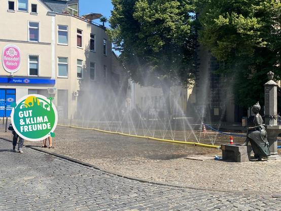 Das Bild zeigt eine gepflasterte Straße in einer Stadt, auf der ein Wasserstrahl aus einem Rohr spritzt, um die Luft zu kühlen. Im Hintergrund sind Gebäude zu sehen, eins davon mit einem Schild "Reissdorf Kölsch" und "Apollo". Einige Bäume mit grünen Blättern stehen links und rechts der Straße. Auf der rechten Seite steht eine Statue einer Frau, die auf einem Sockel sitzt. Im Vordergrund links sind zwei Personen zu sehen, die sich dem Wasserstrahl nähern. Ein Schild mit der Aufschrift "Gut & Klima Freunde" ist im Bild zu sehen. Die Szene wirkt sonnig und lebendig.