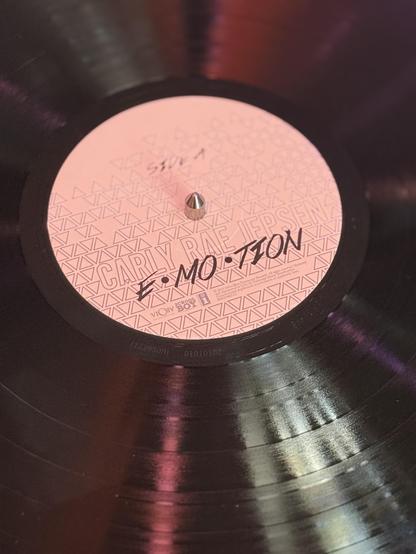 A photograph of the A side of “Emotion” by Carly Rae Jepsen. It is a black vinyl with pink light reflecting off of it. The label is white with a triangle pattern design.