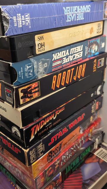 Vhs tapes stacked on top of one another. features movies like last Starfighter, escape from New York, robotjox, temple of doom, Star wars, cky, etc.