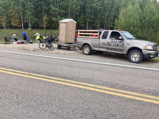 A small pickup truck with a trailier parked on a small roadside stop.  The trailer is very low to the road and has a portable toilet (port-a-potty) mounted on it.  The passenger side door has been painted with a message “Biking to end Cancer, RIDEMN1.ORG”.  A group of cycilists can be seen picture left behind the truck with coolers, camp chairs, and bikes around them.  Dense trees are behind the group in the background.