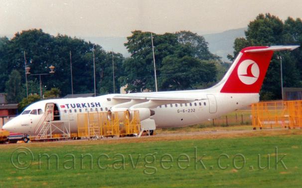 Side view of a high-winged, 4 engined jet airliner parked facing to the left.
The plane is mostly white, with blue "Turkish" titles on the upper forward fuselage, next to a small Turkish flag.
The black registration "G-6-232" is on the lower rear fuselage.
The tail is red, with a white circle containing a stylised red flying bird.
There are several yellow metal frames parked between the camera and the plane, partially obscuring the forward fuselage, as well as some on the right of the image, and a white set of airstairs parked next to the open forward door.
Green grass fills the foreground, with trees lining the background, under grey sky.