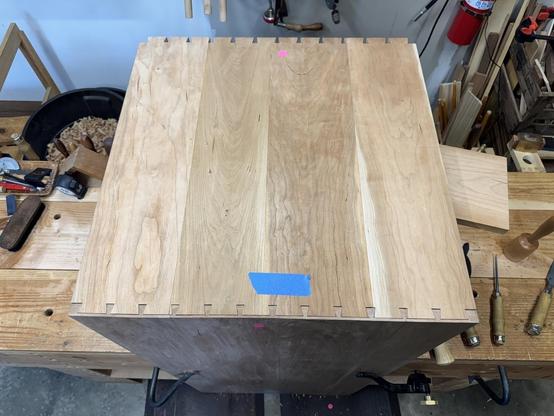 A wooden box with dovetail joints is positioned on a workbench, surrounded by woodworking tools and materials.