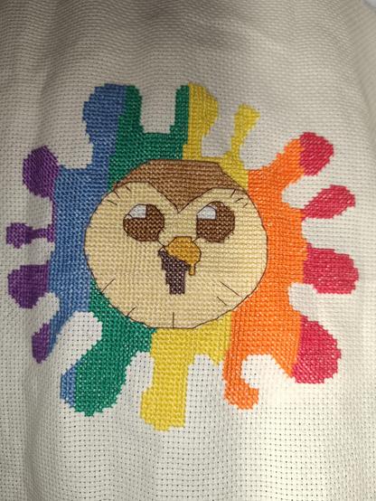Cross stitch featuring face of Hooty from The Owl House cartoon surrounded by a rainbow splat stitched onto cream Aida fabric.