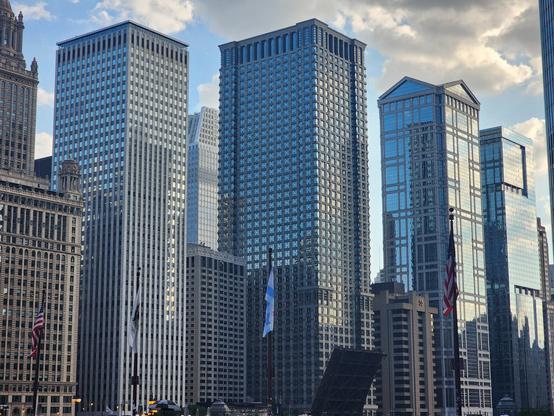 four glass skyscrapers about thirty stories tall standing in an orderly row, towering over smaller buildings between them. their glass surfaces brilliantly reflect each other and the blue sky/clouds.