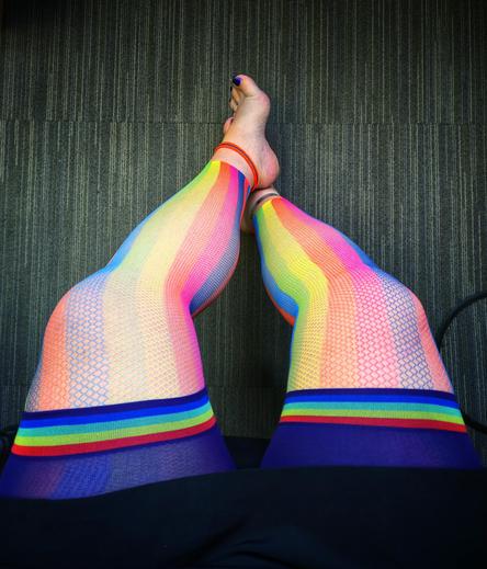 Bare legs crossed playfully on a grey-striped office carpet, wrapped in Snag’s vibrant rainbow fishnet tights from their Pride Collection, each shimmering scale catching the light like candy. Just below, the bold Chub Rub Shorts – Human Nature”peek out, hugging the thighs with a wide rainbow waistband that practically screams power. A neon orange anklet pops against the skin, unapologetically loud, while blue-painted toes flirt with the lens, confidently bare in the workplace. This isn’t just an outfit... it’s a colorful middle finger to dull dress codes and quiet closets.