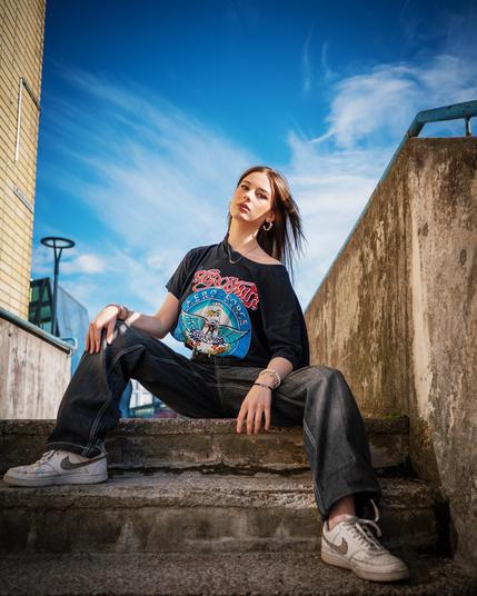Young woman seated on concrete steps with legs apart, wearing a black graphic T-shirt, dark jeans, and white Nike sneakers. Her hair is loose, and she accessorizes with hoop earrings and bracelets. The sky is blue with wispy clouds, and the background includes a brick wall and metal railings.