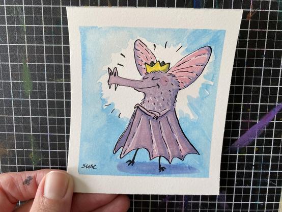 Eine Fledermaus steht Stolz mit Krone und verschränkten Armen da. Farblich ein leichter Lilaton mit grau und großen rosa Ohren.