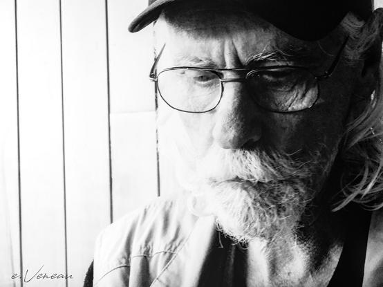 Portrait of an elderly man with a cap, glasses and a white beard. He is looking down.