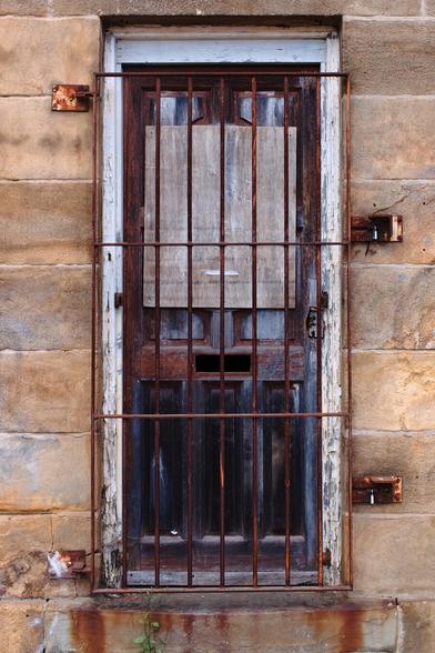 An old wooden door, weathered and darkened with age, is boarded up with a faded panel of plywood and secured behind rusty iron bars. The bars are firmly fixed into the stone wall, which is stained with time and moisture, showing signs of erosion and age. The door’s paint is chipped, especially along the white doorframe, revealing layers beneath. A small rectangular slot sits near the center of the door, hinting it might have once served as a mail slot. Rust drips and a tiny green plant growing from a crack in the stone complete the scene’s sense of abandonment and decay.