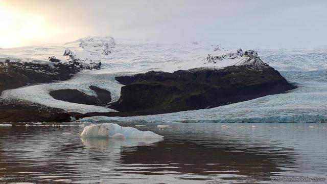 A colour photo of a body of rippled water with icebergs floating in it. Behind it is a mountainside with two glaciers dropping to water level. The sky is grey but the sunlight is glaring on the left. The icebergs are picking up this light on their left faces.
