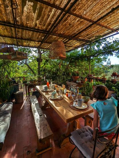 A full, wide-angle shot shows a young child in a light blue top sitting at the end of a long, rustic wooden table, looking out over a lush, green, sunlit balcony filled with potted plants and hanging baskets. The table is set with plates, cups, and a jug of orange juice, suggesting a meal. Above, a slatted, corrugated roof provides shade, and a large, woven light fitting hangs down. A wooden bench runs along one side of the table, and a cushioned chair is on the other. The floor is made of terracotta tiles.