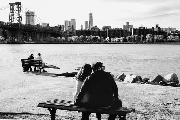 a view onto the williamburg waterfront with the williamsburg bridge tower at the left, 2 couples sit on benches by the brooklyn side of the Hudson, in the distance the newly built WT1 building rises into the center of the frame in manhattan