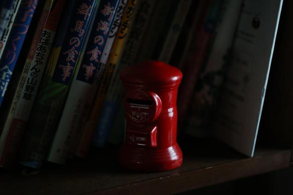 Cookbooks with Japanese titles are lined up at an angle on a dimly lit bookshelf, and in front of them is a red ceramic mailbox-shaped piggy bank, its edges lit by light from outside.