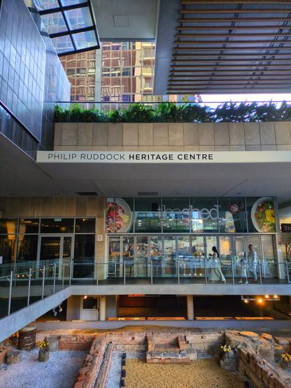 In context. Directly behind the exhibit is a cafe. Above is a sign that says Philip Ruddock Heritage Centre. About two floors up, there's a timber ceiling. Another tall building is visible in the distance.