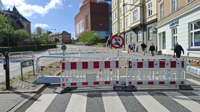 Short version: barriers closing a road to cars and motorcycles. Fuller description: Red and white barriers closing the end of a road to specifically (there's a sign) cars and motorbikes. Beyond, Aros art museum and people walking and cycling as normal on paths to each side. The wide empy car lanes emphasise how much physical public space is handed freely to private car drivers every day, even in a city like Aarhus where many streets do prioritise cyclists and pedestrians.