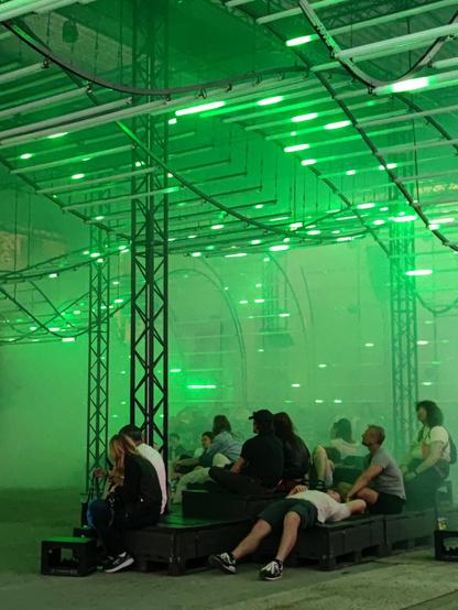Menschen sitzen und liegen auf Kisten unterhalb einer Lichtinstallation. Diese besteht aus Leuchtröhren, die an geschwungenen Schienen an der Decke angebracht sind. In den Röhren sind kleine grüne Lichter zu sehen, die ein wenig wie Glühwürmchen wirken.