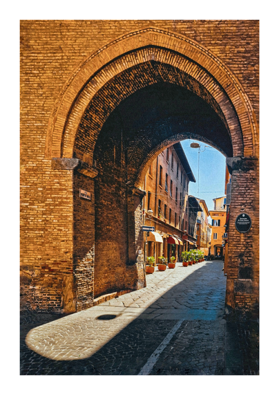 This photo is of an arched city gate leading to a sun-drenched alleyway in Bologna.  The perspective is from beneath the large, brick archway that frames the view down a narrow street lined with old, ochre/yellow-coloured buildings.  Sunlight streams through the archway, creating strong shadows on the cobblestone street.  Plant pots with greenery are visible along the sides of the street. A plaque on the wall of the arch is visible and says "Torresotto di Porta Nuova".